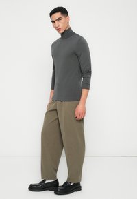 Mand iført en tætsiddende grå turtleneck, løse olivengrønne bukser, hvide sokker og sorte loafers, står mod en hvid væg.