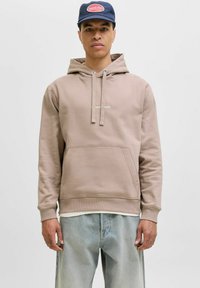 Sudadera beige hecha de material suave, con un bolsillo frontal, capucha con cordón y puños acanalados. Lleva puesta con jeans de color azul claro y una gorra navy.