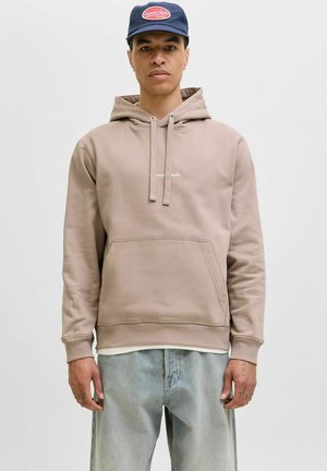 Beige Kapuzenpullover aus weichem Material mit einer Fronttasche, Kordelzug-Kapuze und gerippten Bündchen. Getragen mit hellblauen Jeans und einer marineblauen Mütze.