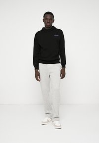 Svart hettegenser med frontlomme, kombinert med lysegrå joggebukser. Modell som har på seg hvite joggesko med beige detaljer.