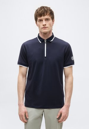 Bărbat tânăr purtând un tricou polo cu mâneci scurte, de culoare albastru închis, cu margini albe și guler cu fermoar, asortat cu pantaloni bej deschis.