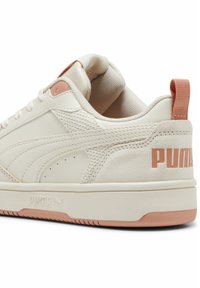 Bež tenisice s glatkim kožnim gornjim dijelom, teksturiranim panelima i svijetloružičastim potplatom. Imaju odvažno "PUMA" brendiranje sa strane.