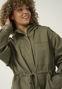 Jachetă cu glugă de culoare verde măslinie, cu talie strânsă, închidere cu fermoar, două buzunare pe piept și manșete elastice. Țesătură moale și texturată.