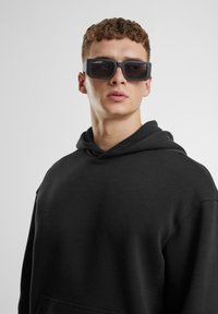 Schwarzer Kapuzenpullover mit lockerer Passform, ausgestattet mit einer vorderen Tasche. Das Model trägt rechteckige, dunkle Sonnenbrillen mit dicker schwarzer Fassung.