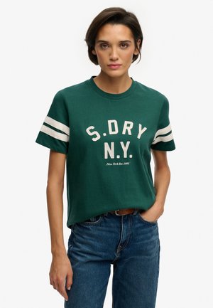 Kvinde iført en grøn kortærmet skjorte med hvide striber på ærmerne og teksten "S.DRY N.Y.", kombineret med blå jeans og et brunt bælte.