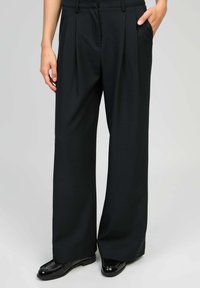 Femme portant un pantalon large noir et des mocassins en cuir verni noirs, se tenant avec une main dans la poche sur un fond gris clair.