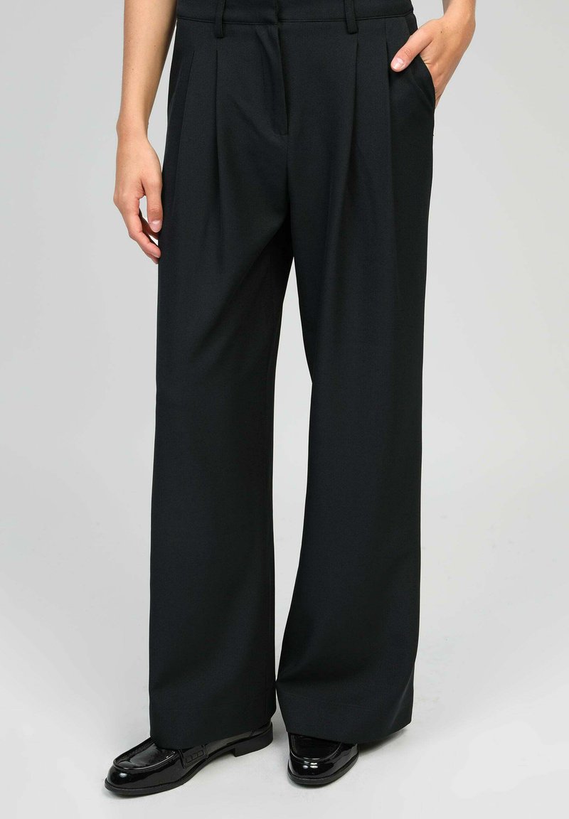 Femme portant un pantalon large noir et des mocassins en cuir verni noirs, se tenant avec une main dans la poche sur un fond gris clair.