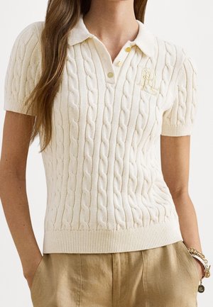 Femme portant un polo crème à manches courtes en maille torsadée avec un logo brodé et un pantalon beige, les mains dans les poches, avec des bracelets au poignet.