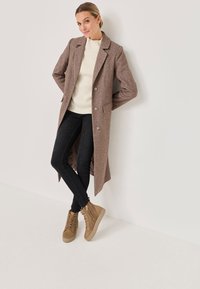 Brauner Wollmantel mit taillierter Silhouette, geknöpftem Vorderteil und Seitentaschen, kombiniert mit schwarzen Jeans und hellbraunen Schnürstiefeln.