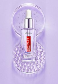 Bouteille en verre transparent de sérum d'acide hyaluronique avec un compte-gouttes blanc, ornée d'une étiquette rouge sur une assiette circulaire à texture bulleuse, le tout sur un fond violet.