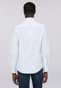 Biała koszula z długim rękawem, o zaokrąglonym dole i zapinanych mankietach, wykonana z gładkiego materiału. Widok z tyłu pokazuje klasyczny krój kołnierzyka.