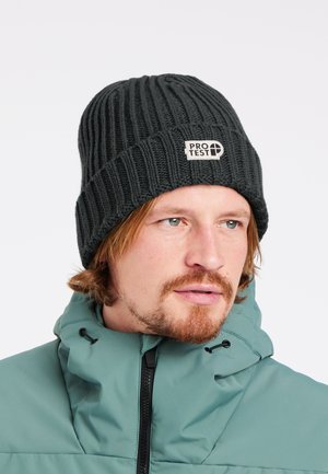 Man met lichte baard en snor, draagt een donker geribde muts met het label "PROTEST" en een teal winterjas met een hoge kraag.