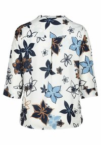 Blouse blanche à manches trois-quarts, présentant de grands imprimés floraux bleus, noirs et marron ainsi qu’un pli central au dos pour un meilleur ajustement.