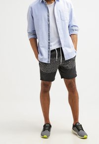 Black swim shorts with a circular pattern, paired with a light blue button-down shirt and gray athletic shoes with neon accents.
