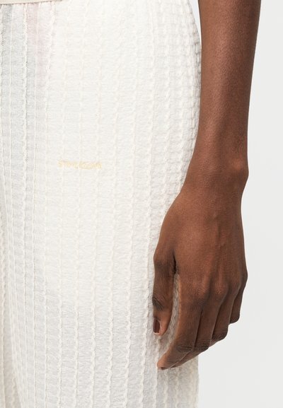 Σκούροχρωμο χέρι που ακουμπάει δίπλα στο ισχίο, φοράει λευκό υφασμάτινο ένδυμα με λεπτά κάθετα σχέδια και μικρό χρυσό λογότυπο.