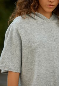 Szürke kapucnis felső, puha, texturált anyagból. Rövid, széles ujjakkal és laza szabással. Minimalista és egyszerű kialakítás.