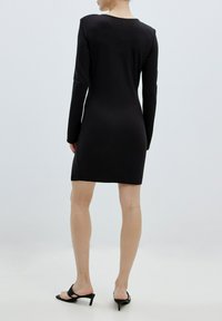 Schwarzes langärmliges Kleid mit einer figurschmeichelnden Silhouette, glatter Textur, hohem Kragen und seitlichen Schlitzen. Kombiniert mit schwarzen Absatzsandalen.