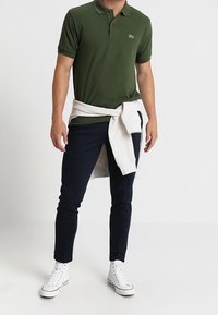 Grön pikétröja, marinblå slim fit-byxor, vita sneakers och en ljusgrå tröja knuten runt midjan. Inga synliga mönster.