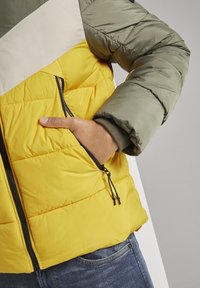 Giacca imbottita con blocchi di colore giallo e verde oliva, caratterizzata da una striscia beige, tasca laterale con zip e polsini elasticizzati. Tessuto testurizzato.