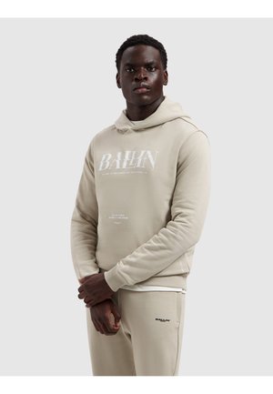 Man staat met gevouwen handen, draagt een beige hoodie en bijpassende trainingsbroek met "BALLIN"-logo tegen een witte achtergrond.