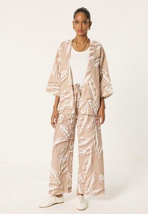 Mujer con pantalones beige de pierna ancha y kimono a juego con patrones blancos de hojas, camisa blanca, joyas doradas y zapatillas blancas, de pie quieta.