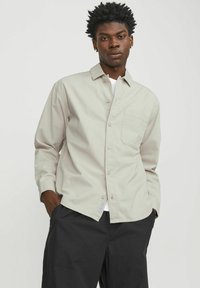 Chemise boutonnée gris clair avec des manches longues, une poche poitrine et une coupe décontractée. Portée par-dessus un T-shirt blanc et associée à un pantalon sombre.