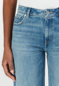 Jeans de denim en azul claro con una cintura alta, cinco bolsillos y una sutil textura desgastada. Presenta un cierre frontal con botón de metal y cremallera.