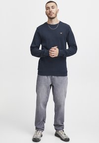 Temno moder sweatshirt z okroglim ovratnikom in rebrastimi manšetami, v kombinaciji s svetlo sivo obleko in sivimi čevlji z teksturiranim vzorcem.