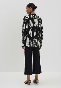 Blusa bianca e nera con motivo a foglie, maniche lunghe e colletto, abbinata a pantaloni neri larghi. La vista posteriore enfatizza il design e la vestibilità.