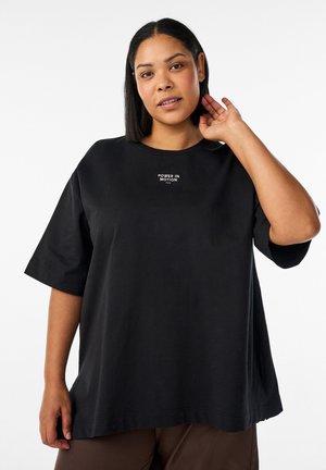 Kvinde med glat sort hår iført en løs sort T-shirt med teksten "Power in Motion", poserer med den ene hånd på nakken.