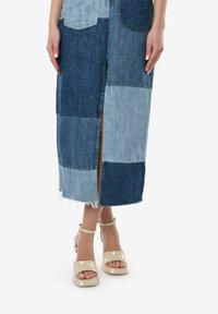 Spódnica z denimu w stylu patchwork z różnorodnymi odcieniami niebieskiego, z rozcięciem z przodu, surowym wykończeniem i zestawiona z beżowymi, lakierowanymi sandałami na koturnie.
