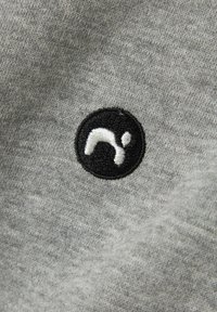 Grauer Stoff mit einer weichen Textur, der ein kreisförmiges schwarzes gesticktes Logo mit weißen Akzenten zeigt. Nahaufnahme der Materialdetails.