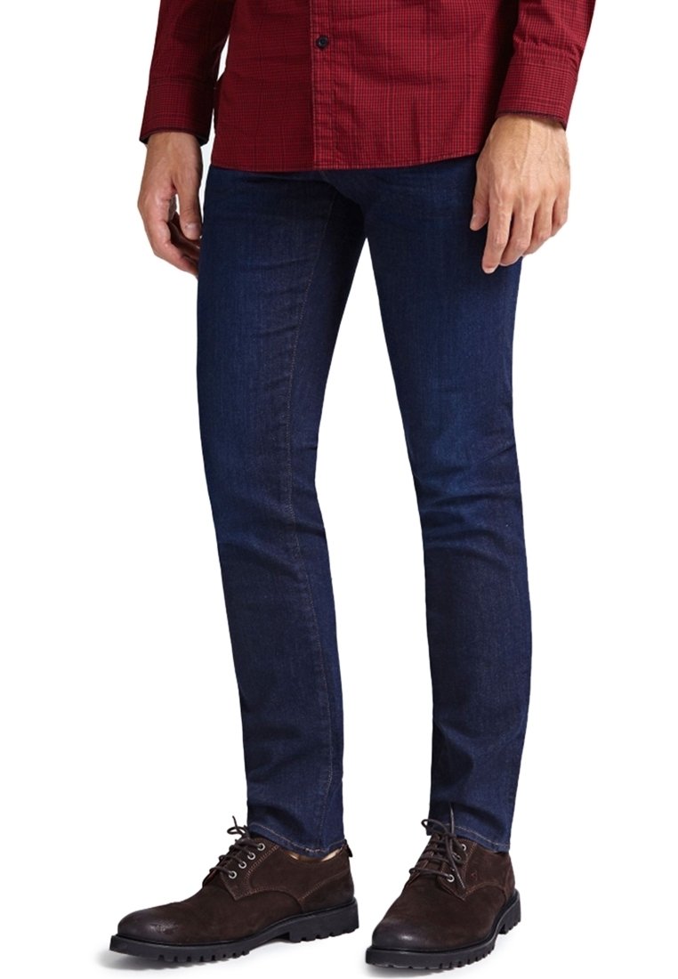 Homme portant un jean slim bleu foncé, des chaussures à lacets marron foncé et une chemise à manches longues à carreaux rouges, debout devant un fond blanc.