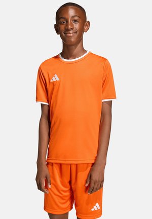 Boy standing and smiling, wearing an orange Adidas sports outfit with white trim on sleeves and Adidas logos on chest and shorts.