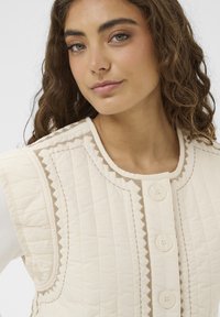 Prošívaná krémová vesta s kulatým výstřihem, doplněná zapínáním na knoflíky vpředu, cikcakovým lemováním a dvěma velkými knoflíky. Měkká textura a zdobená prošitými vzory.