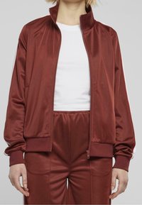 Maroon tracksuit jacket with a zip front, high collar, and white side stripes. Made from smooth, lightweight fabric with ribbed cuffs.
