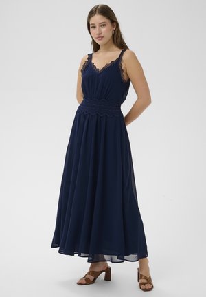 Młoda kobieta stojąca, ubrana w granatową, bezrękawnikową suknię maxi z koronkową lamówką oraz brązowe sandały na obcasie, na białym tle.