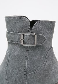 Bottine en daim gris avec une bride décorative et une boucle rectangulaire argentée, coutures détaillées et bord supérieur légèrement plié.