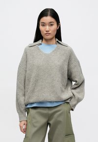 Helehall hall kootud kampsun V-kaeluse ja terava kraega, kantud sinise särgi kohal. Laisaltistuv ja tekstuuriga kangas, kombineeritud roheliste pükstega.