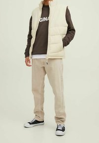 Gilet imbottito beige sopra un felpa marrone con il testo "ORIGINAL", abbinato a pantaloni beige chiaro e sneakers nere. Sfondo neutro.