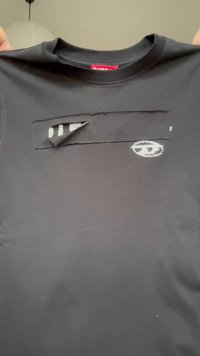 Melns T-krekls, kurā ir daļēji nolobīta taisnstūra forma melna plāksteris, atklājot zem tā baltiem burtiem tekstu un nelielu baltu ovālu logotipu uz krūtīm.