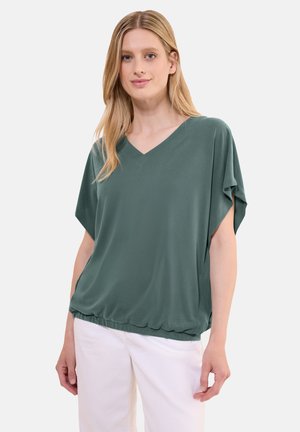 Top verde a maniche corte con scollo a V realizzato in tessuto liscio, caratterizzato da una vestibilità rilassata e orlo elastico, indossato sopra pantaloni bianchi.