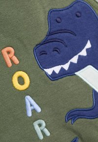 Der grüne Sweatshirt-Stoff zeigt einen blauen Dinosaurier mit weißen Zähnen und bunten, gestickten Buchstaben, die "ROAR" in Orange, Gelb, Hellblau und Grün buchstabieren.