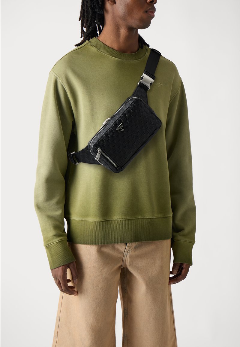 Black quilted crossbody bag with a triangular logo, silver zippers, and adjustable strap, worn over an olive green sweatshirt and beige pants.