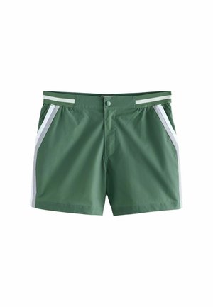 Grønne herre badebukser med hvide sidelister, knaplukning foran, båret af en skulderløs mand, der står ved vandet.