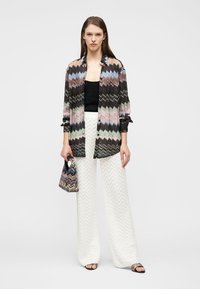 Femme portant une chemise à motif zigzag multicolore, un haut noir, un pantalon blanc à jambes larges, tenant un petit sac assorti, debout sur un fond blanc.