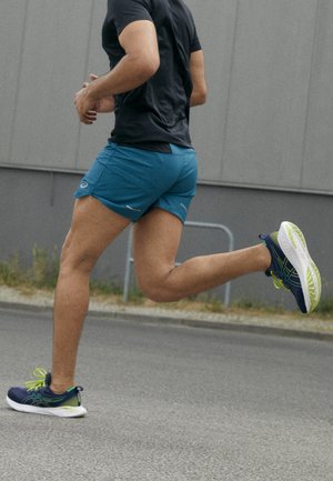 Abbigliamento da corsa maschile con una maglietta nera aderente a manica corta e pantaloni corti turchesi. Le scarpe sono blu navy con accenti verdi e un design leggero.