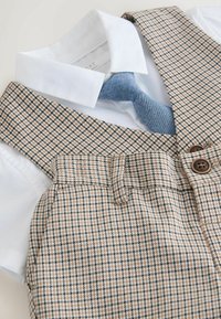 Camisa blanca con cuello y corbata azul claro combinada con tirantes beige y marrón con estampado de pata de gallo y pantalones de talle alto a juego con botones.