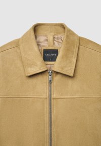 Hellbraune Wildlederjacke mit Reißverschluss, Stehkragen und dezenten Nahtdetails über der Brust. Das Etikett zeigt "CALLIOPE" in Schwarz.