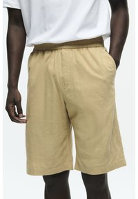 Pantaloni corti beige in cotone con vita elastica, tasche laterali e una vestibilità rilassata. Il tessuto ha un aspetto morbido e strutturato.